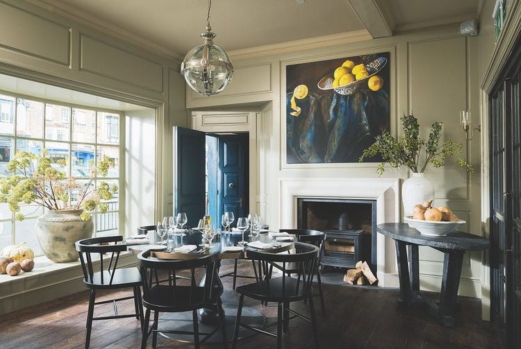 A well-lit, elegant dining room with a chandelier, large windows, and a wall painting, featuring tables, chairs, and decorative items.